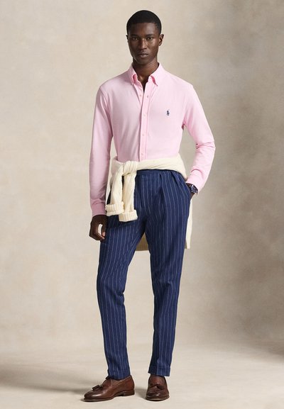Light pink button-down shirt, navy pinstriped trousers, cream sweater tied at the waist, brown leather shoes, and a watch on the left wrist.