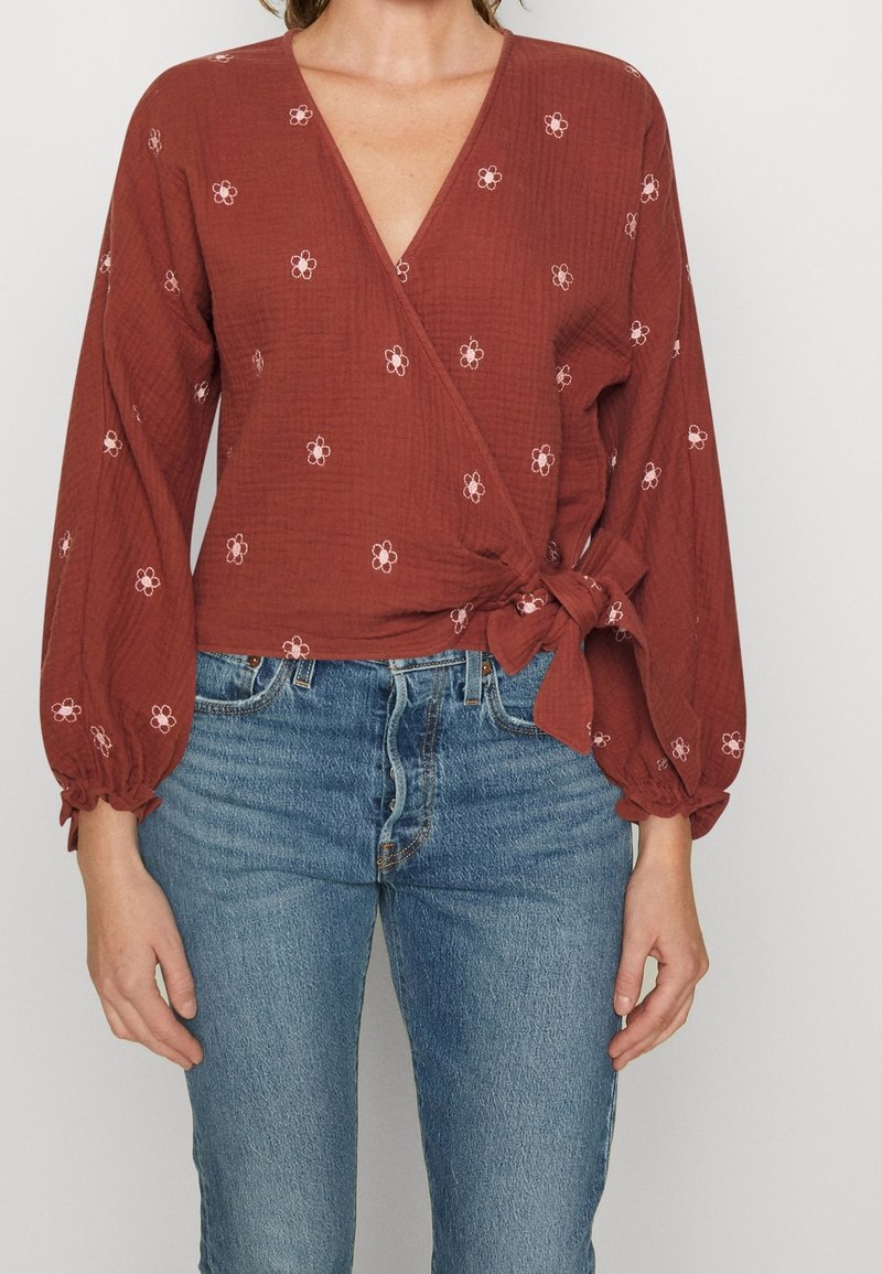 Rostfarbene Wickelbluse mit tiefem V-Ausschnitt, langen Puffärmeln und bestickten rosa Blumen; am Bund gebunden. Kombiniert mit blauen Jeans.