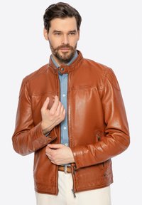 Giacca di pelle marrone con colletto alto, chiusura a zip, tasche laterali e texture liscia, indossata sopra una camicia azzurra chiara.