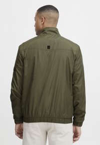Olive green lightweight jacket with a stand collar, elastic cuffs, and hem. Features a black label detail on the back. Smooth, matte texture.