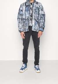 Osoba stojí v modro-bíle vzorované bundě, černých kalhotách a modrých teniskách, na jednoduchém bílém pozadí.