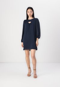 Vestido mini azul-marinho com mangas bufantes longas, detalhe no decote em forma de olho-chave, tecido texturizado e um corte solto. Combinado com sandálias de tiras em tom bege.