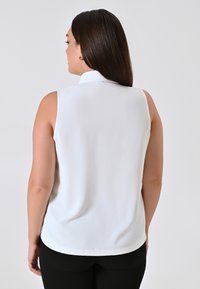 Blusa bianca senza maniche con colletto, tessuto morbido e taglio all'altezza dei fianchi, abbinata a pantaloni aderenti neri.