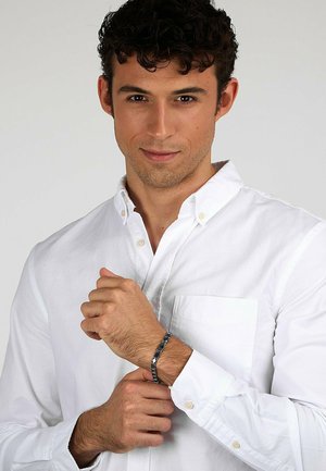 Weißes Hemd mit Knöpfen, Modell hält das Handgelenk. Das Armband besteht aus dunkelblauen und silbernen Gliedern, hat eine glatte Textur. Schlichtes Design, modernes Aussehen.