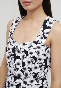Kvinna som bär en ärmlös topp i svart och vitt med breda axelband och abstrakt blommönster, som visar halsringning och överkropp.