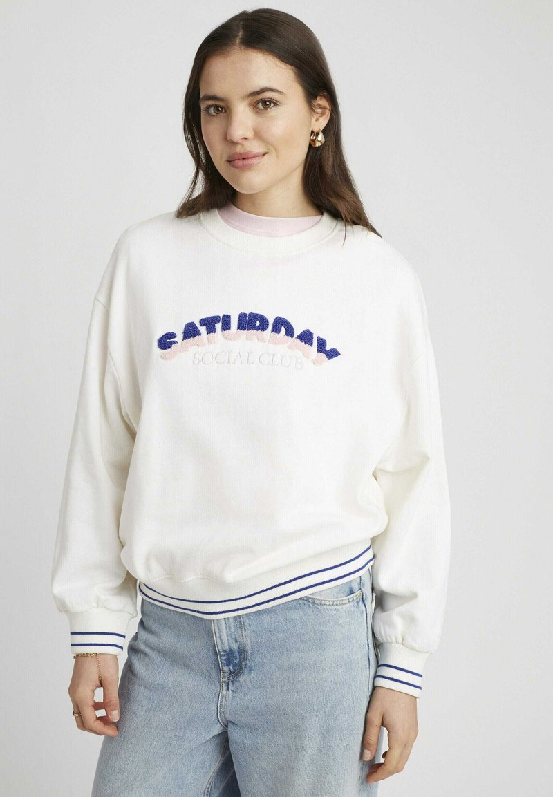 Sweat-shirt blanc avec broderie "SATURDAY SOCIAL CLUB" en rose et bleu. Présente des poignets et un ourlet côtelés avec des rayures bleu marine. Coupe décontractée.