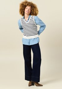 Gestreifter ärmelloser Strickwesten in Weiß und Marineblau über einem hellblauen Knopfhemd, kombiniert mit dunkelblauen weiten Jeans und braunen Absatzstiefeln.