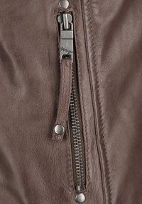 Detail einer braunen Lederjacke mit einem silbernen Reißverschluss und einem Zugriegel, sichtbaren Nähten und einem metallischen Druckknopfakzent.