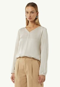 Lichtbeige blouse met een V-hals, lange mouwen met elastische manchetten en een schijnbare naad aan de voorkant. De stof lijkt glad en lichtgewicht.