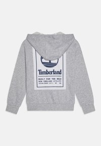 Siva dukserica s kapuljačom s bijelim pravokutnim grafičkim dizajnom s plavim Timberland logotipom i tekstom na leđima. Mekani materijal i ležeran kroj.