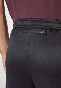 Calças atléticas pretas com acabamento texturizado, apresentando um cós elástico e um bolso com fecho de correr na parte de trás para armazenamento.