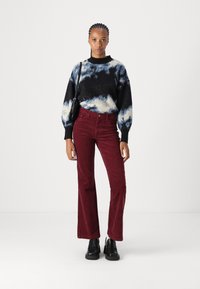 Svetr z pleteniny s černým a modrým tie-dye vzorem, červené kordové kalhoty s širokými nohavicemi a černé robustní boty, s malou černou kabelkou přehozenou přes rameno.