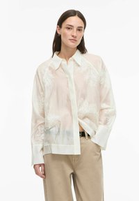 Chemise blanche en tissu transparent avec des motifs floraux brodés, un col avec rabat, et des manches longues, associée à un pantalon beige clair.