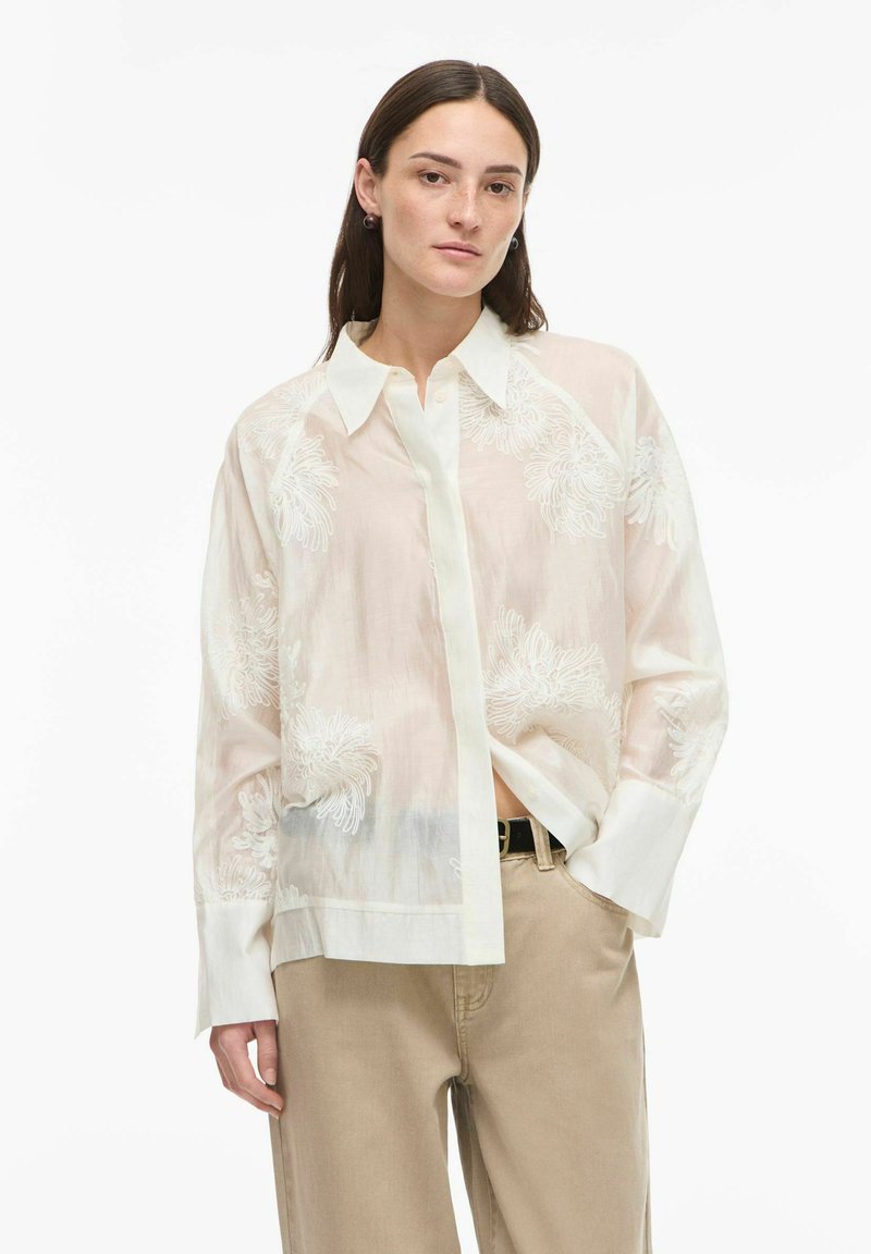 Chemise blanche en tissu transparent avec des motifs floraux brodés, un col avec rabat, et des manches longues, associée à un pantalon beige clair.