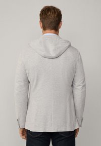 Grå stickad blazer med huva, som har långa ärmar, en slät textur och en skräddarsydd passform. Anmärkningsvärda detaljer inkluderar knäppningar och en ventilerad rygg.