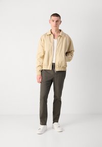 Beige bomberjack met een zachte textuur, voorzien van een ritssluiting aan de voorkant, elastische manchetten en zakken, gecombineerd met olijfkleurige broeken en witte sneakers.