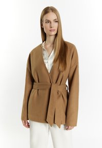 Cardigan de culoare cămilă, cu un croi înfășurat și talie legată, având mâneci lungi și o textură moale, purtat peste o bluză din satin alb.