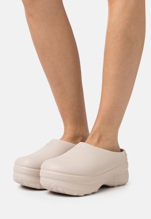 Sabots à plateforme beige à enfiler, avec une semelle épaisse, portés pieds nus, présentant un design minimaliste et un embossage discret du logo Adidas.