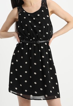 Schwarzes, ärmelloses Kleid mit großen weißen Polka-Punkten, tailliertem Bund und einem schmalen schwarzen Gürtel. Verfügt über eine halbtransparente Spitzenverzierung an den Schultern.