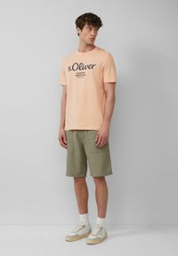 Perzikkleurig katoen T-shirt met marineblauwe bedrukking, gecombineerd met olijfgroene shorts. Accessoires zijn witte sokken en lichtgrijze sneakers met beige zolen.