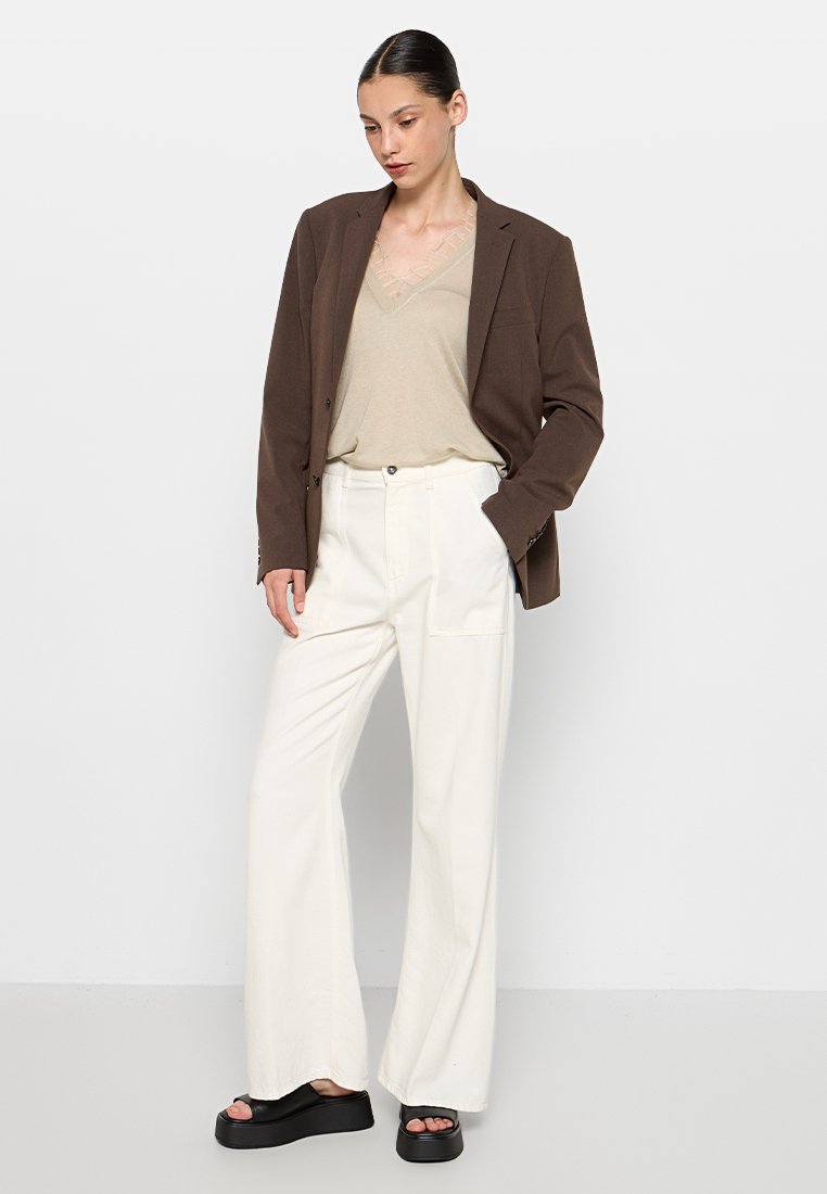 Bruine blazer over een lichtbeige V-hals top, gecombineerd met wijde witte broek en zwarte platform sandaal. Soepele texturen, moderne pasvorm.