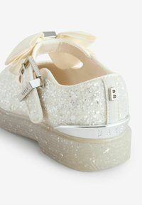 Sneaker bianco glitterato con tomaia testurizzata, un grande fiocco crema in accentuazione, hardware argentato e suola in gomma trasparente. Etichetta del marchio "BAKER" in evidenza.