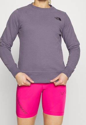 Person iført en lilla langærmet sweatshirt med The North Face-logo og lyserøde løbeshorts med Dynafit-logo.