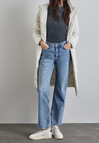 Kvinna i ljusbeige vadderad kappa, mörkgrå ribbad topp, hög midja blå jeans och vita sneakers står med händerna i fickorna.