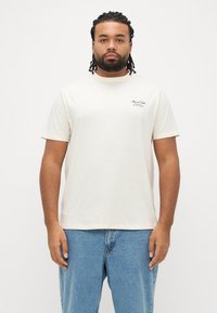 Lysebeige bomuld t-shirt med korte ærmer; har et subtilt sort logo på brystet, rund halsudskæring og løs pasform.