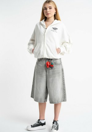 Młoda kobieta ubrana w biały bluzy z kapturem na zamek, szare długie dżinsowe szorty z breloczkiem w kształcie czerwonego serca, białe falbankowe skarpetki i czarne trampki stoi.