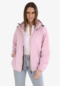 Veste de pluie rose clair avec capuche, fermeture éclair à l'avant et poches latérales. Possède une fermeture à glissière contrastante noire et jaune. Matériel lisse et léger.