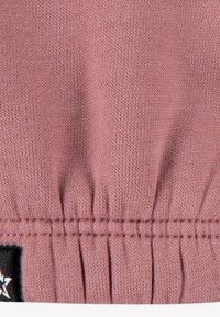 Tissu tricoté rose avec un motif texturé, présentant des détails froncés à l'ourlet et une petite étiquette de logo noire visible sur le côté.