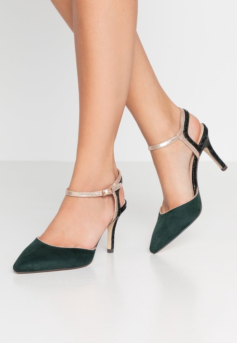 Chaussures à talons hauts en daim vert avec des pointes effilées, une bride métallique dorée et des accents en vernis noir au niveau du talon et de la cheville, dans un design élégant.