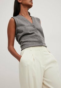 Grijze mouwloze gebreide top met een v-hals en vijf knopen, gecombineerd met crèmekleurige high-waisted broek met plooien en zijzakken.