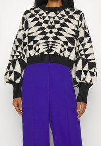 Pull en tricot noir et crème à motifs, avec un design court, des manches larges et un ourlet côtelé, associé à un pantalon violet taille haute.