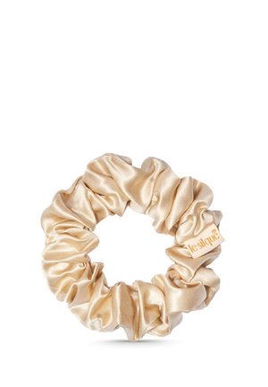 Chouchou en satin beige avec une petite étiquette en tissu portant la mention "leslique,", disposé en plis circulaires rassemblés sur un fond blanc.