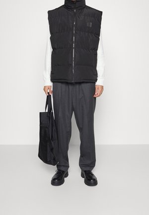 Czarna pikowana kamizelka z wysokim kołnierzem, zapinana na zamek, noszona na białej koszuli i szarych szerokich spodniach, dopełniona czarną torbą typu tote.