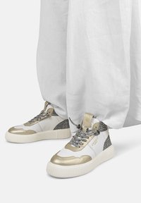 Białe sneakersy z metalicznymi złotymi akcentami, czarne detale w panterkę, szare teksturowane sznurowadła oraz masywna biała podeszwa.