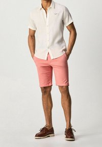 Biała lniana koszula z krótkim rękawem z guzikami, w połączeniu z różowymi szortami i brązowymi skórzanymi butami typu boat z białą podeszwą.