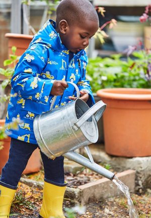 Niño con un chubasquero azul con patrones de vehículos amarillos, sosteniendo una regadera metálica, de pie en un jardín con macetas de terracota.