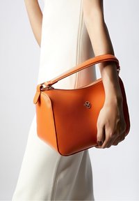 Oranssi nahkainen käsilaukku, jossa on jämäkkä muoto ja lyhyt, irrotettava hihna. Korostaa kultaa logoa ja sileää pintaa.