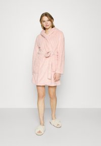 Pink, fluffy bathrobe with a hood, tie belt, and two front pockets. Paired with cream-colored open-toe slippers. Soft texture throughout.