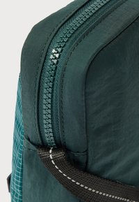 Türkizkék szövet hátizsák sarka sima cipzárral, megerősített varrással és fekete szíjjal, amelyen fehér díszítések találhatók.