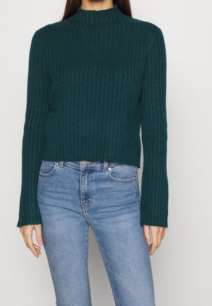 Dark teal ribbed sweater with a high neck and long sleeves, paired with light blue fitted jeans. Sweater features a cropped design and textured fabric.