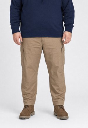 Man draagt beige cargobroek, bruine veterlaarzen en een donkerblauw shirt met lange mouwen, staand tegen een effen achtergrond.