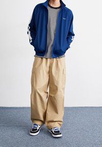 Henkilö, jolla on päällään sininen vetoketjullinen takki, musta-valkoiraitainen paita, vaaleat cargo-housut ja musta-valko-sinisiä lenkkareita, seisoo matolla.