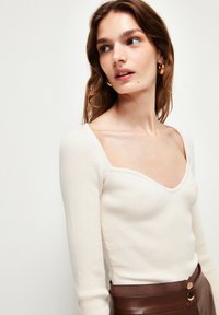 Un haut beige clair à rayures, à manches longues et à décolleté carré profond, associé à une jupe haute marron foncé ornée de boutons dorés.