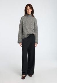 Grauer Fischgrat-Strickpullover mit hohem Kragen, kombiniert mit schwarzen weiten Hosen mit asymmetrischem Design und burgunderfarbenen Slippern.