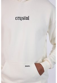 Sudadera blanca con texto impreso en negro "crystal" en el pecho y "dreams." en el bolsillo frontal. Cuenta con un bolsillo tipo canguro y puños acanalados.