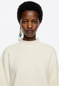 Cremfarbener, strukturierter Pullover mit hohem Kragen. Das Model trägt ein auffälliges Ohr hängen mit Perlen und Charms, das dem Look Details verleiht.